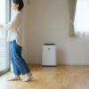 空気清浄機が稼働する明るい日本のリビングで、窓を開けて換気をする女性。窓から入る新鮮な空気と、室内の空気が入れ替わる様子を描写した、アスペクト比16:9の横長画像。